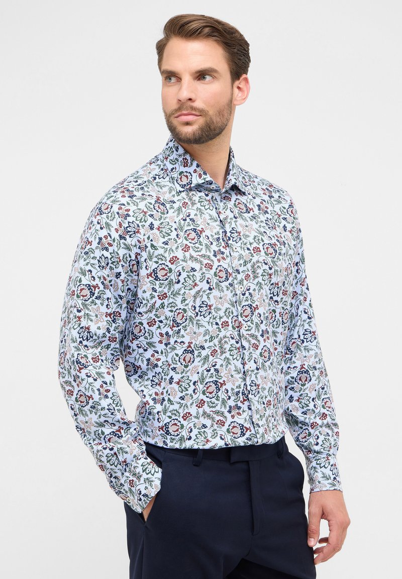 Camicia bianca con bottoni, caratterizzata da un motivo floreale in tonalità di rosso, blu e verde. Maniche lunghe e colletto strutturato. Abbinata a pantaloni scuri.