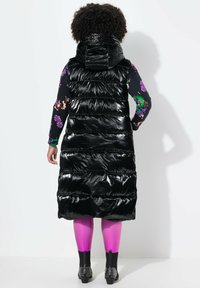 Schwarze Steppweste mit Kapuze, glänzender Textur und horizontalem gestepptem Design, kombiniert mit leuchtend pinkfarbenen Leggings und genagelten schwarzen Stiefeln.