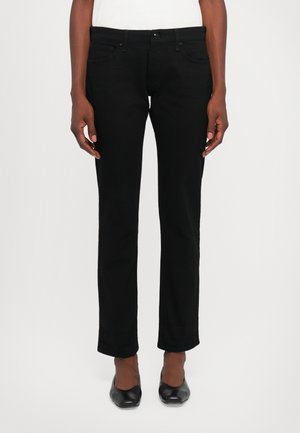 Person wearing straight-cut black pants, black pointed-toe shoes, and a white top, standing against a white background.
