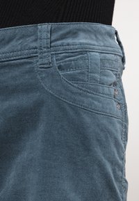 Pantalones de algodón azul con una textura suave, que cuentan con un pequeño bolsillo frontal, costuras decorativas y dos botones metálicos en el lateral.