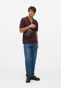 Czarna torebka crossbody z teksturowanym wzorem, noszona na burgundowej koszulce z krótkim rękawem i niebieskich dżinsach, stojąca na białym tle.