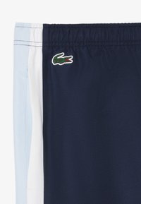 Marineblauwe shorts met een gestructureerde weving, voorzien van witte en lichtblauwe zijpanelen. Een klein Lacoste krokodillenlogo is op de voorkant gestikt.