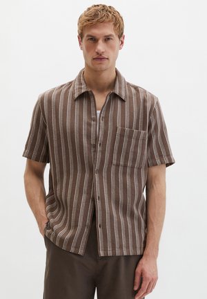 Férfi rövid ujjú, barna ingben, amelyen függőleges fehér csíkok és egy mellzseb található, egyik kezét a zsebében tartva áll.
