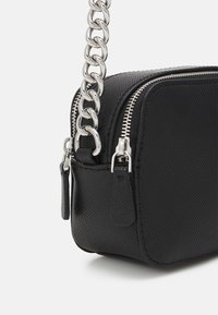 Černá texturovaná kožená kabelka přes rameno se stříbrným zipem a krátkým masivním stříbrným řetízkem, zobrazená v detailu na bílém pozadí.
