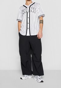 Maglia da baseball bianca a righe con dettagli neri, con bottoni e la scritta 'KANI', abbinata a pantaloni neri larghi e sneakers bianche.