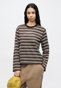 Fiatal nő hosszú fekete hajjal, barna csíkos hosszú ujjú ingben és bézs nadrágban, egyik kezében sárga sapkát tartva.