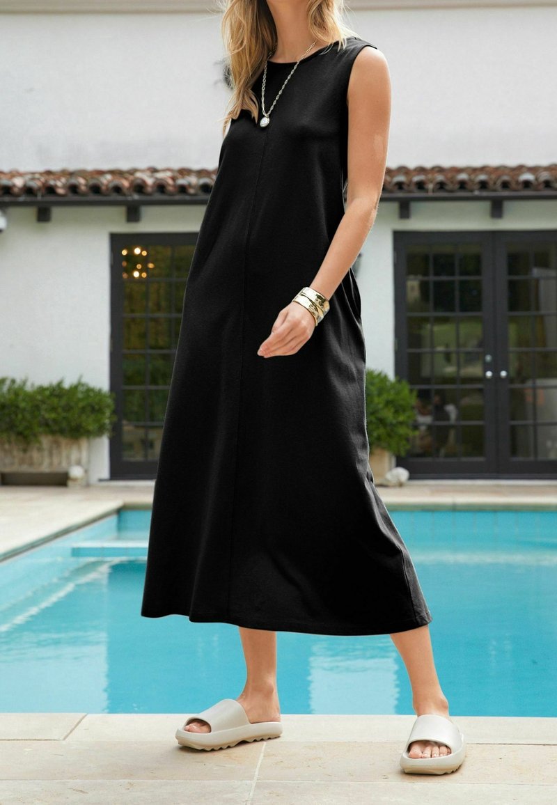 Robe maxi sans manches noire avec une coupe décontractée, dotée d'une couture centrale. Associée à des sandales ouvertes à bout découvert de couleur beige clair. Cadre extérieur autour de la piscine.