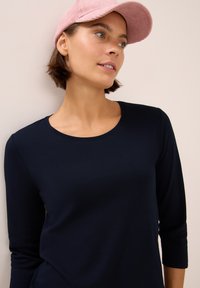 Junge Frau mit kurzem braunem Haar, die eine pinke Mütze und ein navyblaues Langarmshirt trägt und leicht zur Seite schaut.