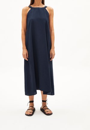 Mujer con un vestido midi azul marino sin mangas con un cuello alto y sandalias planas negras con tiras, de pie frente a un fondo blanco.