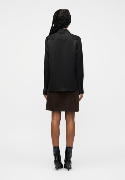 Γυναίκα με πλεγμένα μαλλιά, που φοράει μαύρο πουκάμισο με μακριά μανίκια, σκουρόχρωμη καφέ φούστα και μαύρα μποτάκια με τακούνι, στέκεται με την πλάτη στραμμένη.