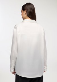 Camicia di seta bianca con colletto, maniche lunghe e polsini con bottoni. Tessuto liscio e orlo arrotondato. Nessun motivo visibile.