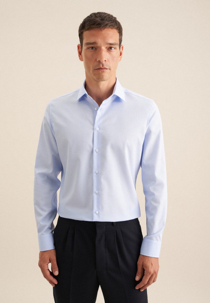 Camicia a maniche lunghe azzurro chiaro con colletto button-down e bottoni bianchi; realizzata in tessuto liscio; abbinata a pantaloni scuri.