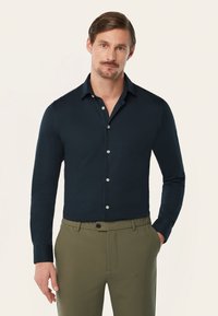 Marineblauwe blouse met lange mouwen, een klassieke kraag, knoopsluiting aan de voorkant en zilveren knopen. Gecombineerd met olijfgroene broeken met zijzakken.