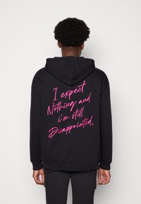 Svart hoodie med en stor rosa handskriven text på baksidan som säger: "Jag förväntar mig ingenting och jag är fortfarande besviken." Tillverkad av bomullsmaterial.