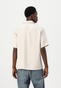 Lys beige kortermet skjorte med vertikale striper, spiss krage og rett snitt; brukt sammen med blå denimjeans.