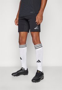 Zwarte sportshorts met een reflecterend logo, gecombineerd met witte sokken met drie zwarte strepen en zwarte voetbalschoenen.