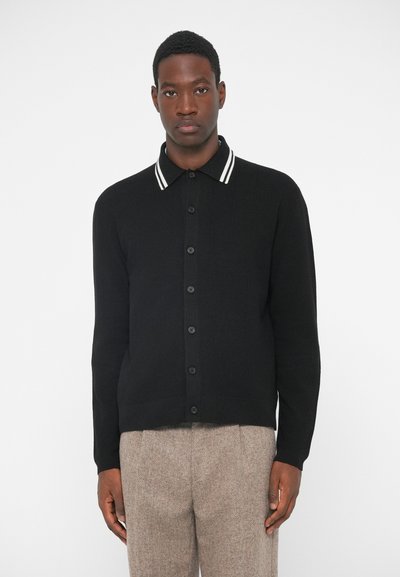Cardigan lavorato a maglia nero con un motivo testurizzato, bottoni neri e colletto a righe bianche in contrasto. Indossato con pantaloni beige a fantasia.
