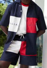 Camicia a maniche corte con bottoni, composta da sezioni blu navy, bianche e rosse con logo. Abbinata a pantaloncini sportivi blu navy e rossi con coulisse.