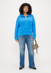 Pulover tricotat de culoare bleu royal, cu guler cu fermoar, având un model texturat de sârmă. Asortat cu blugi din denim albastru și o geantă mică cu imprimeu de zebră.