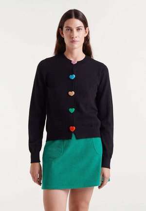 Cardigan nero con bottoni a forma di cuore in rosa, blu, verde, marrone, arancione e rosso; indossato su una gonna in corduroy verde con tasche.