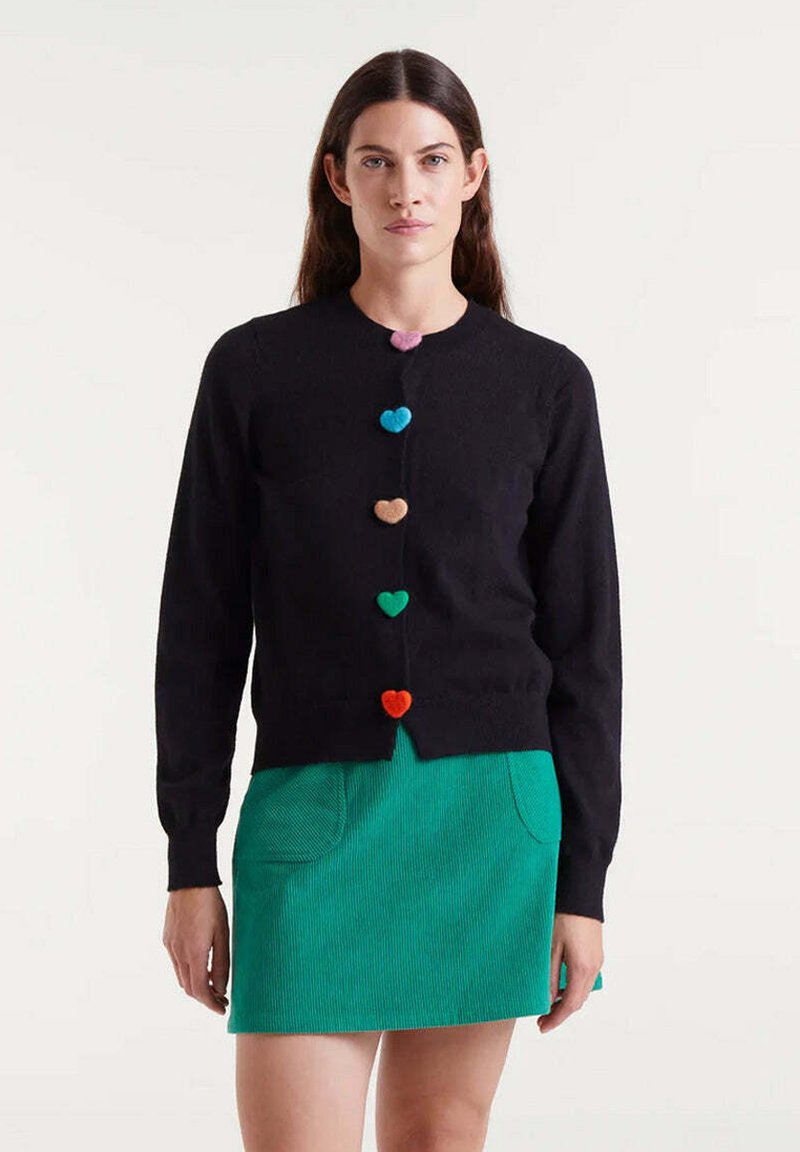 Schwarze Strickjacke mit herzförmigen Knöpfen in Pink, Blau, Grün, Braun, Orange und Rot; getragen über einem grünen Cordrock mit Taschen.