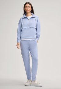 Donna che indossa una felpa blu chiaro con zip e pantaloni jogging abbinati con sneakers bianche, in piedi davanti a uno sfondo neutro.