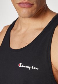 Crna majica bez rukava s izrezom u obliku okruglog izreza i vidljivim šavovima. Sadrži mali bijeli logo "Champion" s crvenim akcentima.