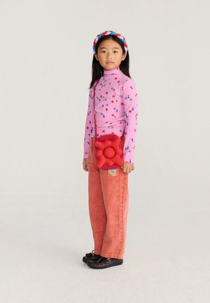 Jong meisje met lang zwart haar, gekleed in een roze bloemencoltrui, rode broek, gevlochten hoofdband en draagt een rode bloemvormige tas.