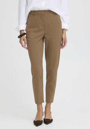 Kobieta ubrana w brązowe, dopasowane spodnie do kostek, białą koszulę na guziki z podwiniętymi rękawami i ciemnobrązowe szpilki z noskiem w szpic, stoi na białym tle.