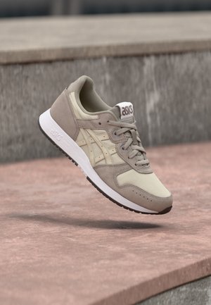 Beiger und hellbrauner ASICS-Sneaker mit weißer Sohle, schwebend über einer steinernen Oberfläche im Freien.