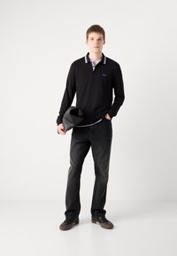 Polo de manga longa preto com colarinho riscado em roxo, combinado com jeans escuros e ténis negros. O modelo segura uma bolsa preta.