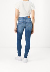 Jeans ajustados de denim azul medio, diseño de cintura alta, con dos bolsillos traseros, combinados con una camiseta blanca y zapatillas blancas.