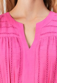 Närbild av en rosa blus med vertikala spetsränder och en V-formad halsringning, bärd av en person med ljus hy och blont hår.