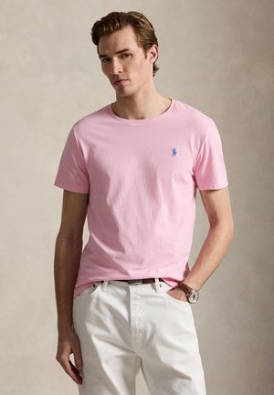 Junger Mann trägt ein hellrosa kurzärmeliges T-Shirt mit blauem Logo und weiße Hose, steht mit einer Hand in der Tasche.