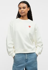 Cream-colored sweatshirt with a round neckline, ribbed cuffs, and hem. Features a small red heart logo on the left chest.