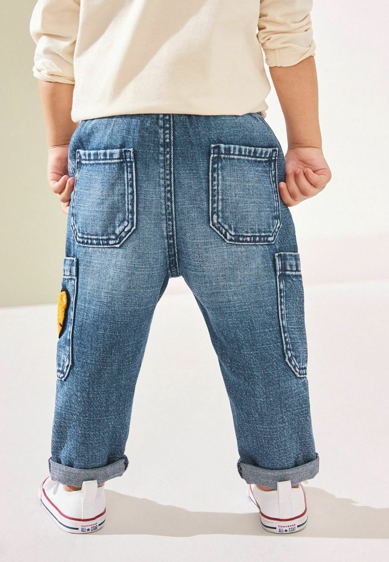Enfant portant un jean bleu retroussé avec des poches arrière et latérales, des baskets blanches et un t-shirt à manches longues beige clair.