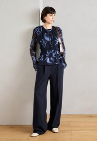 Blouse marine avec des motifs abstraits bleus, design peplum, manches transparentes ; associée à un pantalon large rayé marine et des chaussures de couleur claire.