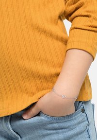 Pulsera de plata con motivos de mariposa y corazón, llevada en una mano dentro del bolsillo de un denim azul claro, junto a un suéter de mostaza texturizado.