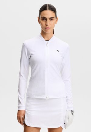 Kobieta o ciemnych włosach ubrana w białą kurtkę na zamek, do tego dopasowaną spódnicę i golfową rękawiczkę na lewej ręce, stojąca na neutralnym tle.