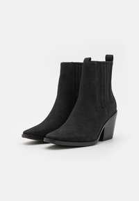 Botas de tobillo de ante negro con punta afilada, tacón bajo y acentos de ribetes verticales en los lados. Presenta una tira en la parte posterior.