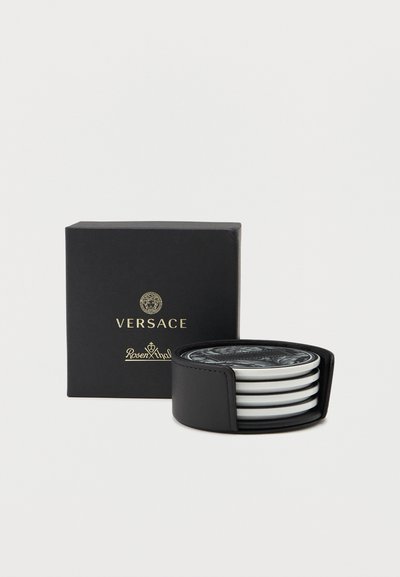 Σετ ασπρόμαυρων υποστηριγμάτων ποτηριών Versace με  σχέδιο Μέδουσας, στοιβαγμένα μέσα σε μαύρη δερμάτινη θήκη, τοποθετημένα δίπλα σε μαύρο κουτί Versace.