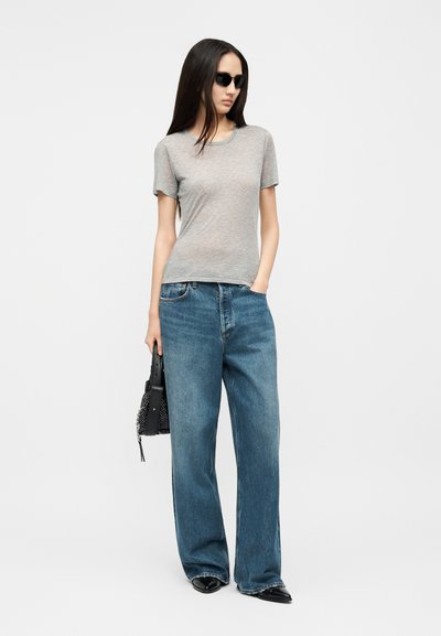 Žena u crnim sunčanim naočalama, sivoj majici, širokim plavim trapericama, crnim šiljastim cipelama, držeći crnu torbu s zakovicama, stoji na bijeloj pozadini.