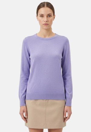 Donna con capelli castani lisci che indossa un maglione a maniche lunghe viola chiaro e una gonna beige, in piedi davanti a uno sfondo bianco semplice.