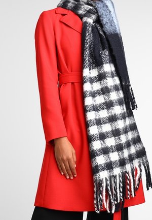 Femme portant un manteau ceinturé rouge vif et une grande écharpe à carreaux noir et blanc avec des franges sur un fond uni.
