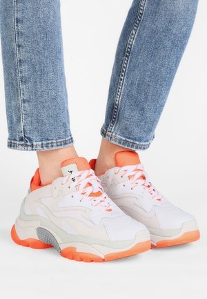 Baskets épaisses blanches et orange clair portées avec un jean bleu, montrant des semelles texturées et un design à lacets.