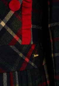 Tkanina w kratę w kolorach ciemnej zieleni, czerwieni i beżu; ozdobiona czerwoną falbanką i złotą metką z logo; miękka tekstura wełny.