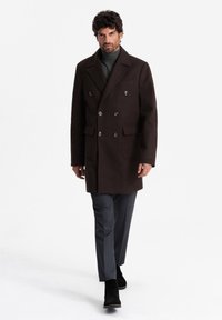 Brown double-breasted coat made of thick wool with notched lapels, two front pockets, and silver buttons, paired with gray trousers and black boots.
