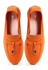 Mocassins en daim orange avec un bout arrondi, un nœud décoratif noué et des accents métalliques. Semelle en caoutchouc de couleur claire. Texture lisse dans l'ensemble.
