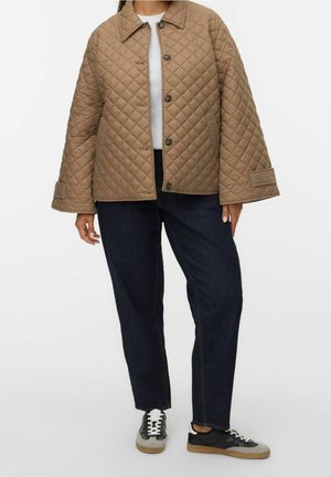 Personne portant une veste matelassée marron à manches larges, un jean foncé, des baskets noires et grises, et un haut blanc sur un fond uni.