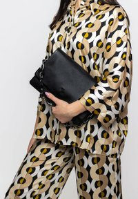 Clutch in pelle nera con tracolla a catena tenuto contro un outfit a fantasia caratterizzato da cerchi nei toni beige, neri e gialli, con finitura in satin.